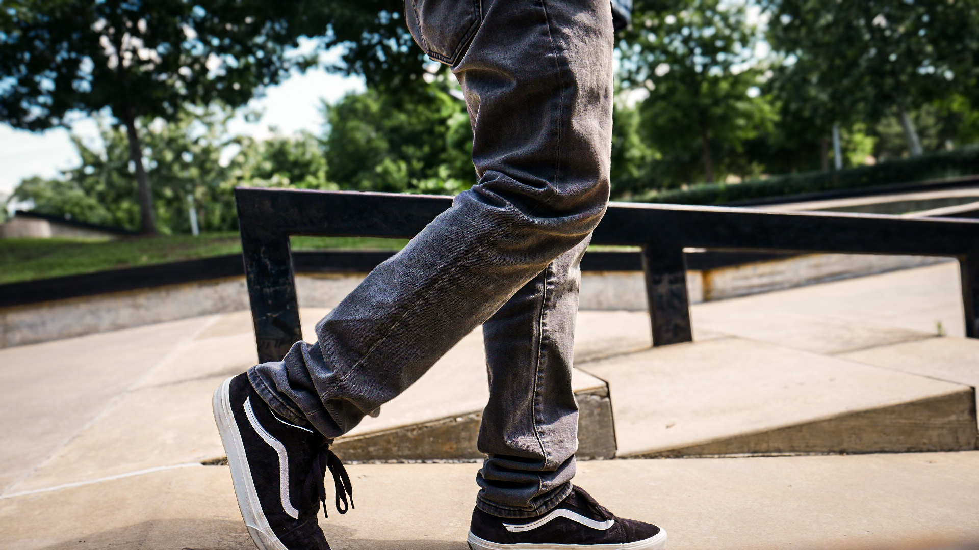 Cinematic street scene: grey jeans and black Vans stepping past a skate-park rail